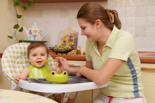 Мама в оливковой футболке, кормит малыша в детском стульчике с ложечки находясь на в кухне