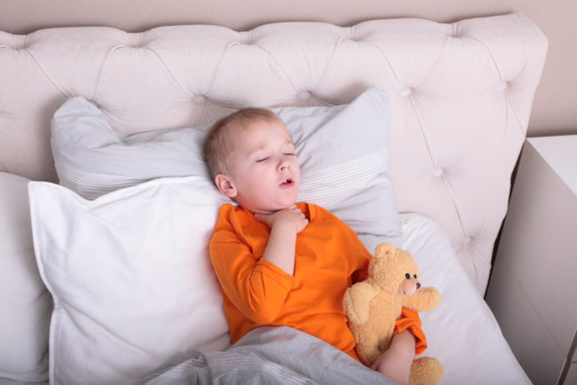 Ребёнок в оранжевой кофте лежит в кровати и держится за горло с игрушкой в руках