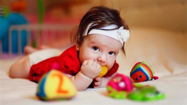 Маленькая девочка с карми глазами и белой повязкой на лбу лежит с игрушкой во рту
