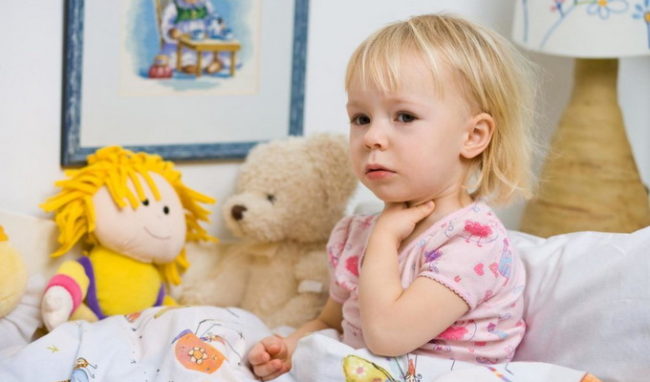Маленькая девочка со светлыми волосами в розовой майке сидит на кровати и держится за горло