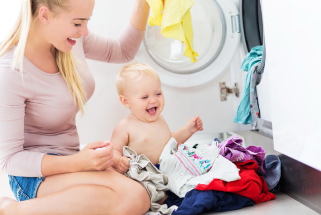 Мама с новорождённым ребёнком сидят с детскими вещами у стиральной машинки