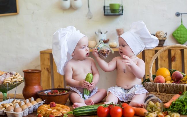 Два малыша в поварских колпаках и сидя среди овощей кормят друг друга