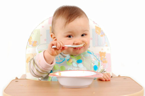 Пятимесячный малыш, сидя в детском стульчике, кушает из тарелки ложечкой