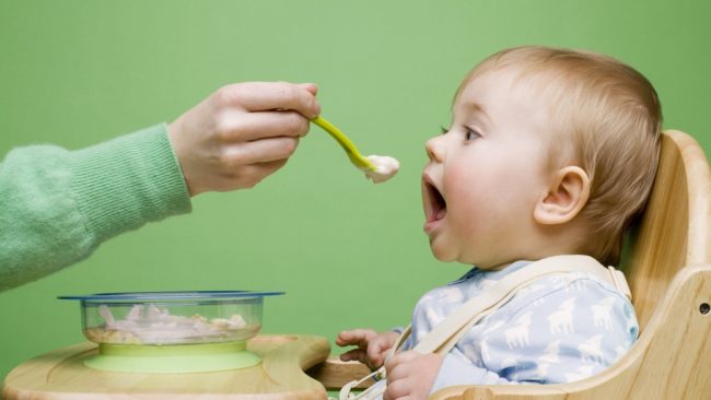 На зеленом фоне пятимесячного малыша кормят с салатовой ложки кашей с прозрачной тарелки