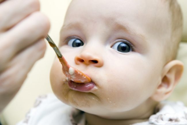 Личико малыша с серыми глазами которого кормят с ложки яблочным пюре.