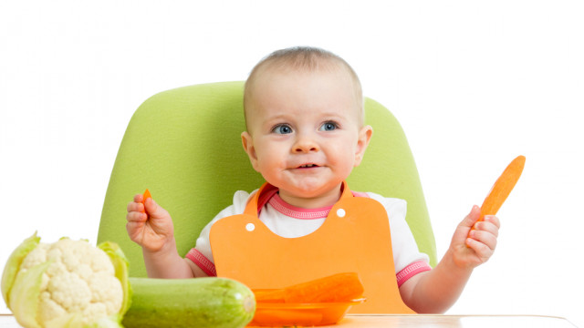 Ребенок сидит в салатовом детском стульчике, перед ним лежит кабачок и морковь