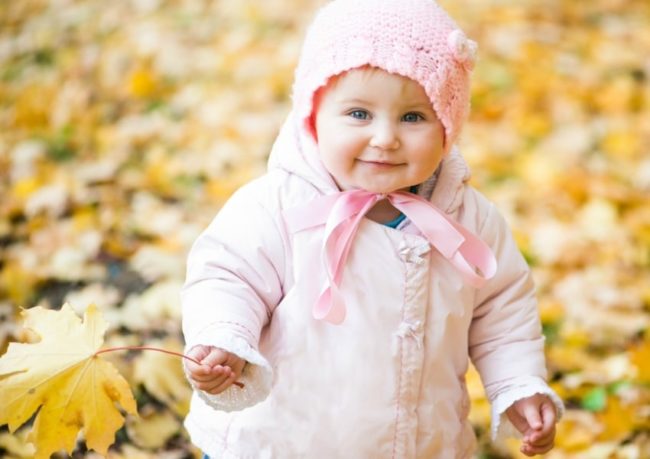 Малышка с голубыми глазами в розовом пальто и розовой шапке держит осенний листок
