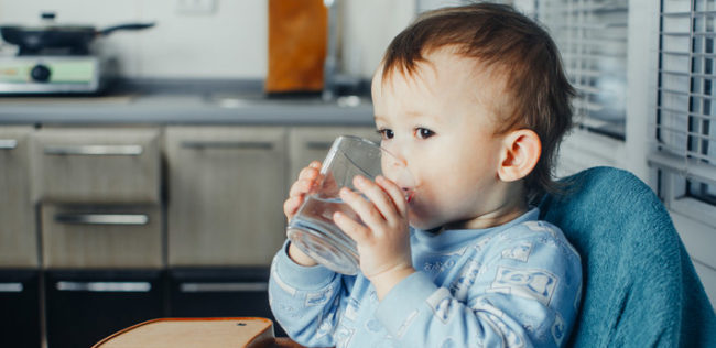 Малыш в голубой кофте держит стакан в руках и пьёт воду.