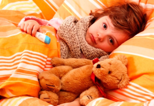 Ребенок с перевязанном шарфом горлом лежит в постели накрытый одеялом и держит термометр в ручке